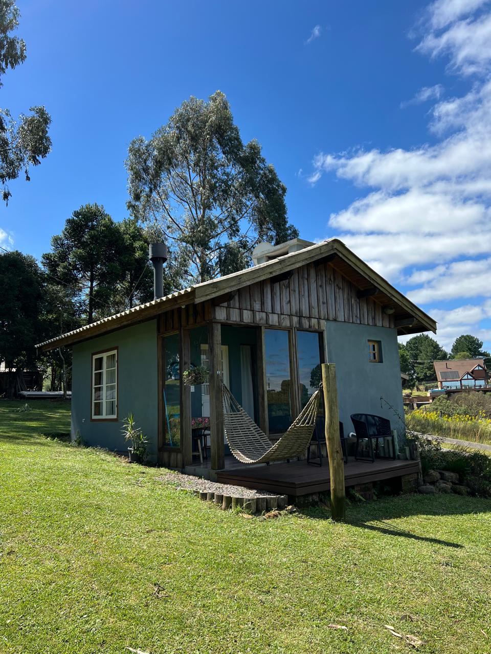 Chalé com varanda, rede e cadeiras, cercado por gramado sob céu azul